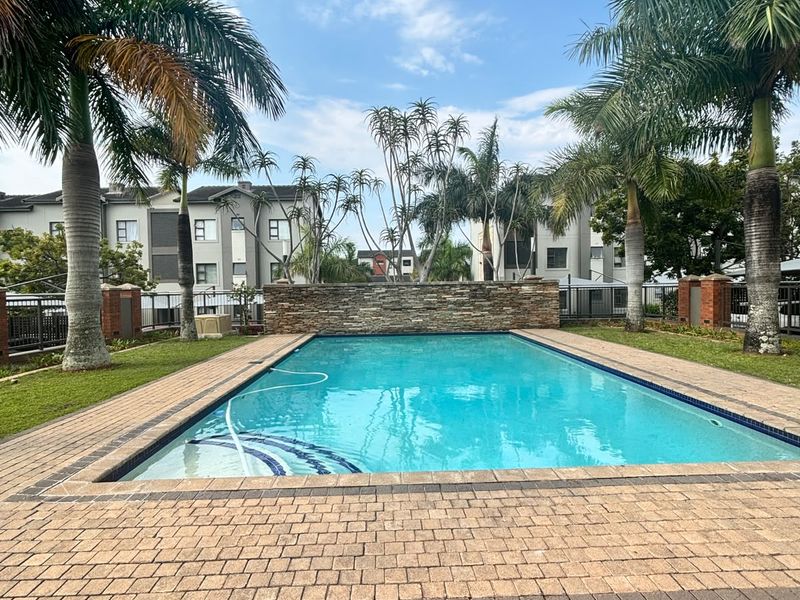 A well-maintained communal swimming pool area featuring a stone accent wall, paved decking, and surrounding landscaped gardens within an apartment complex.