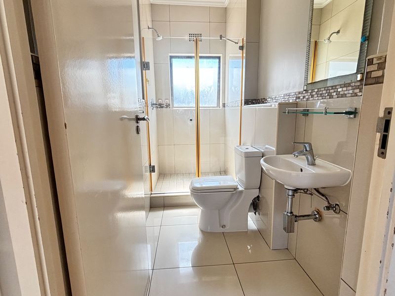 A compact, functional bathroom featuring a shower enclosure, wall-mounted basin, and neutral floor-to-ceiling tiling.
