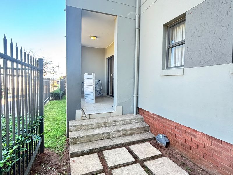 Rear exterior view of the apartment featuring a covered patio, concrete stepping stones, and metal security fencing along the perimeter.
