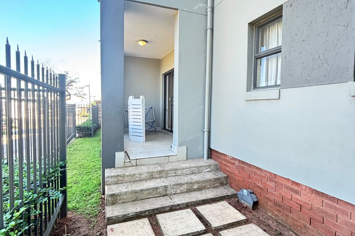 Rear exterior view of the apartment featuring a covered patio, concrete stepping stones, and metal security fencing along the perimeter.
