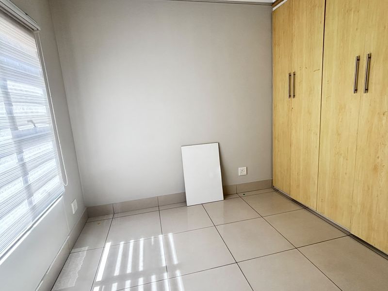 A bright, empty bedroom featuring neutral tiled flooring, built-in wooden cupboards, and modern roller blinds, offering a clean and versatile living space.