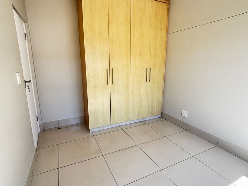 A clean, empty bedroom featuring neutral tiled flooring, a built-in wooden wardrobe, and coved ceiling details, offering a bright and versatile living space.