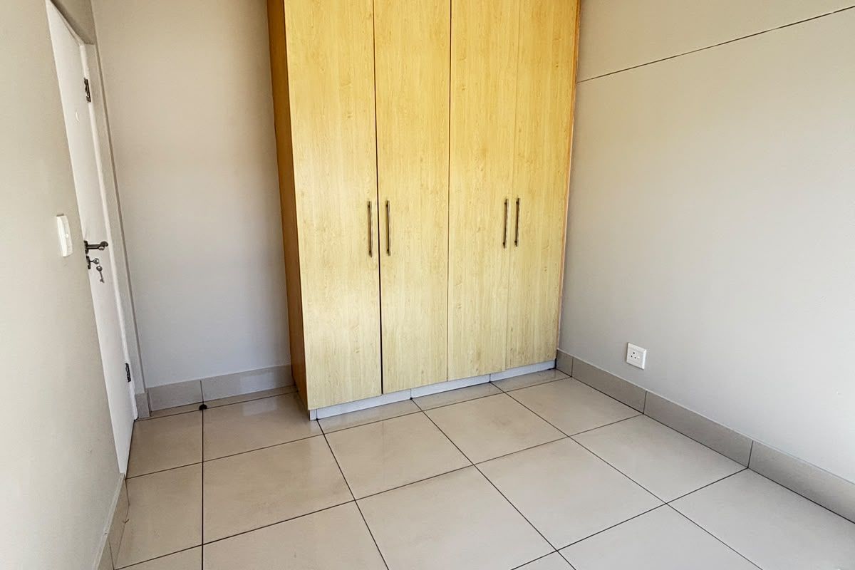 A clean, empty bedroom featuring neutral tiled flooring, a built-in wooden wardrobe, and coved ceiling details, offering a bright and versatile living space.