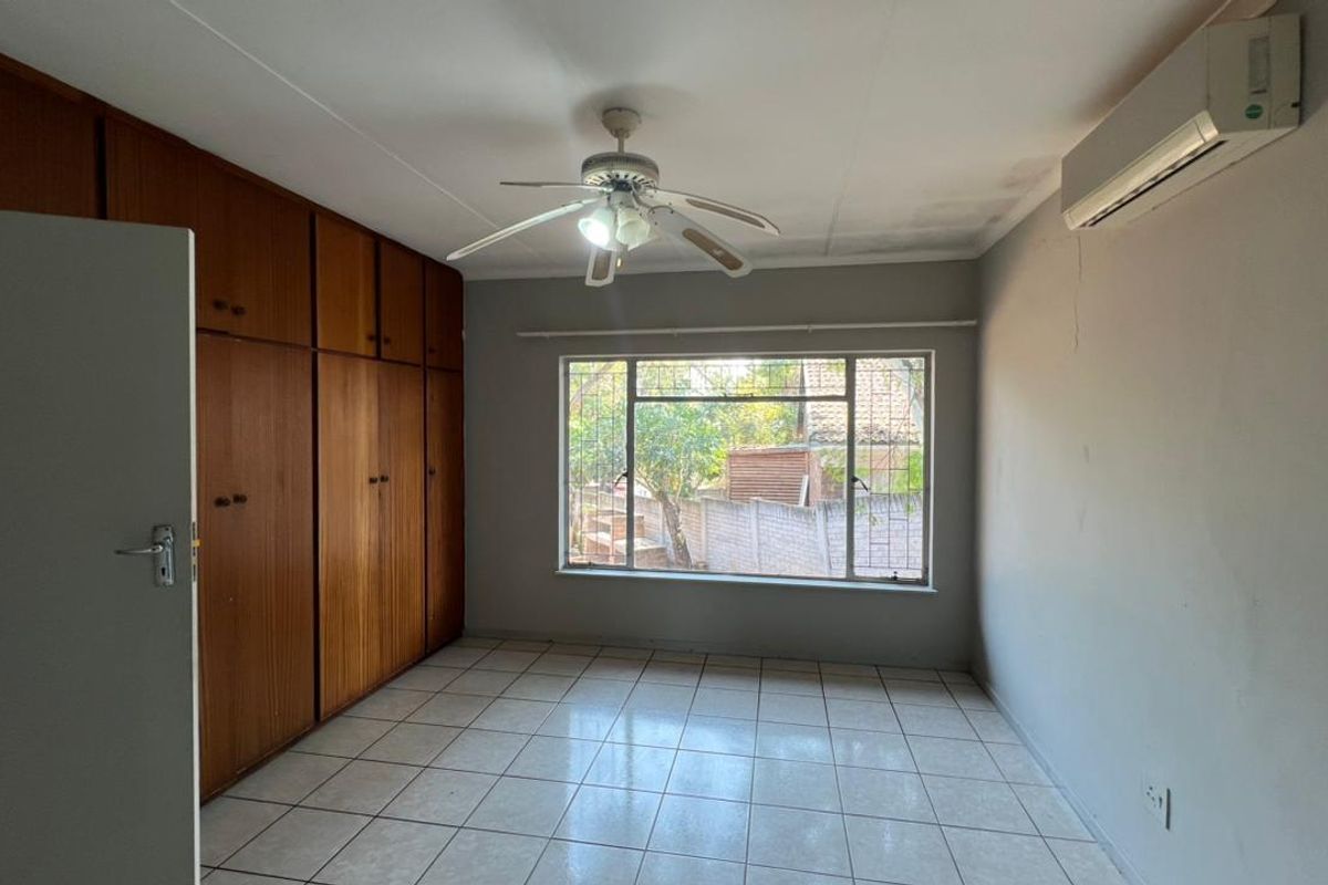 A vacant bedroom featuring built-in wooden cupboards, tiled flooring, a ceiling fan, and a wall-mounted air conditioning unit.