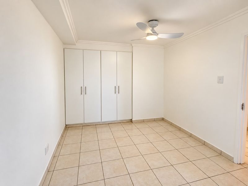 A clean, empty bedroom featuring neutral tiled flooring, built-in cupboards, and a modern ceiling fan, offering a bright and versatile living space.