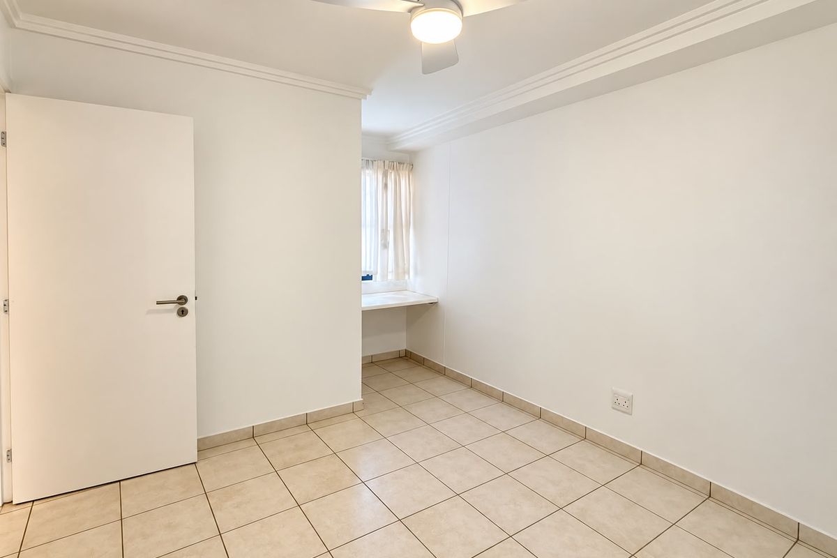A bright, empty bedroom featuring neutral tiled flooring, a ceiling fan, and a built-in desk area, offering a clean and versatile living space.