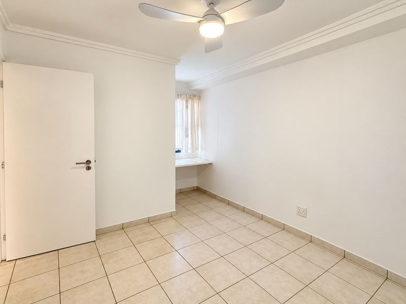 A bright, empty bedroom featuring neutral tiled flooring, a ceiling fan, and a built-in desk area, offering a clean and versatile living space.