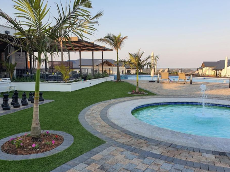 Well-maintained outdoor pool area featuring a central water fountain, paved patio, artificial lawn, and a covered patio space for relaxation.