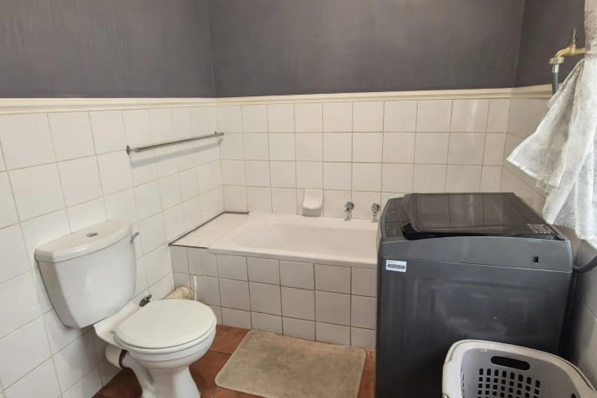A functional bathroom featuring a built-in bathtub, pedestal basin, and toilet, with half-height ceramic wall tiling and a window for natural light.