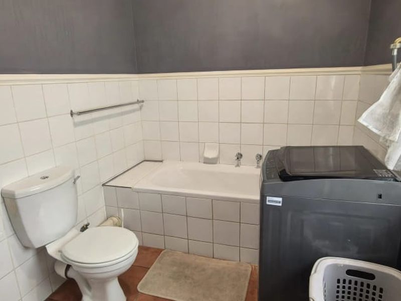 A functional bathroom featuring a built-in bathtub, pedestal basin, and toilet, with half-height ceramic wall tiling and a window for natural light.