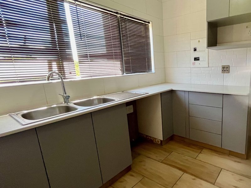 A clean, modern kitchen featuring grey cabinetry, stone countertops, a double sink, and tiled flooring, offering a functional and well-lit workspace.