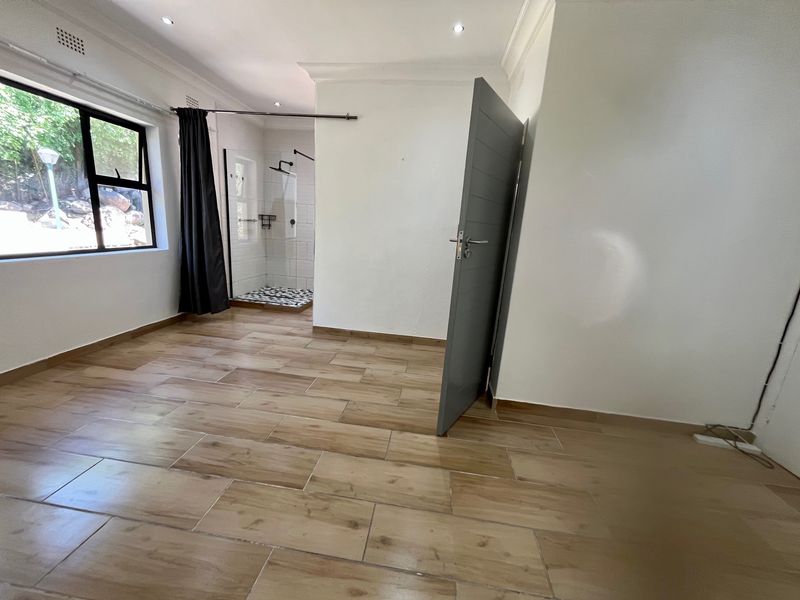 A bright, modern bedroom featuring wood-look tile flooring, recessed lighting, and an integrated en-suite shower with a glass enclosure.
