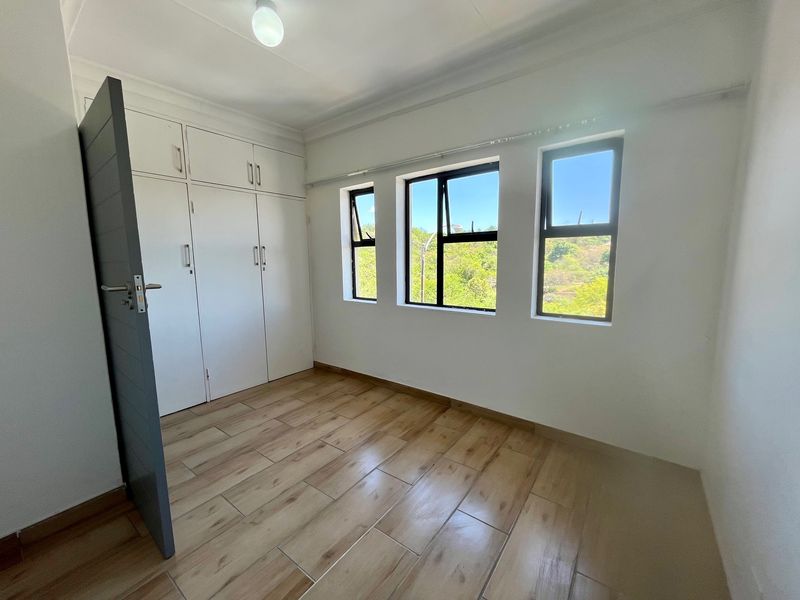 A bright, empty bedroom featuring modern wood-look tiled flooring, built-in cupboards, and large windows providing natural light and scenic views.