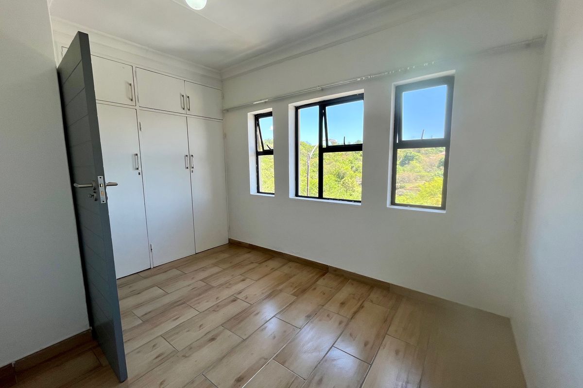 A bright, empty bedroom featuring modern wood-look tiled flooring, built-in cupboards, and large windows providing natural light and scenic views.