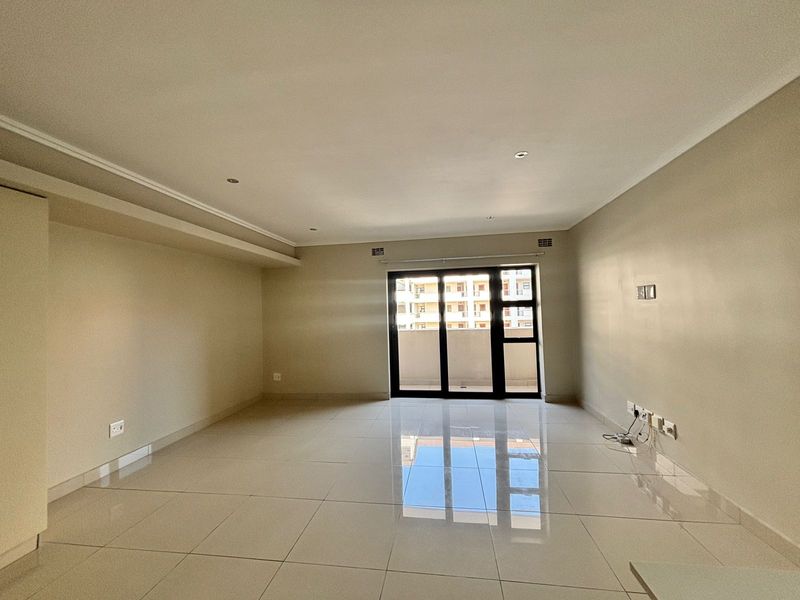 A spacious, empty living room featuring neutral tiled flooring, recessed lighting, and a large sliding glass door providing access to the balcony.