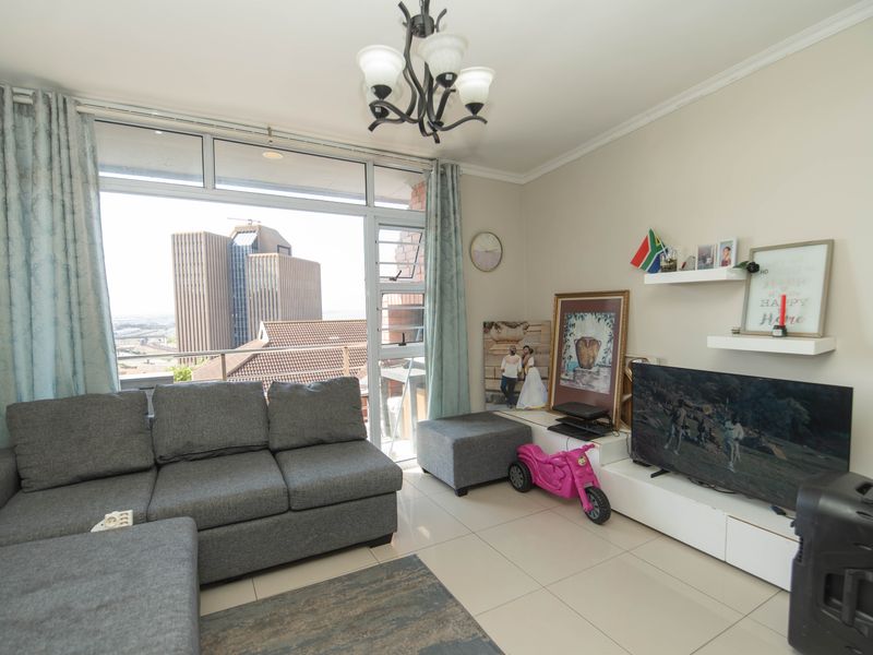A bright living room featuring large windows with city views, tiled flooring, and a neutral color palette, offering a comfortable and well-lit living space.
