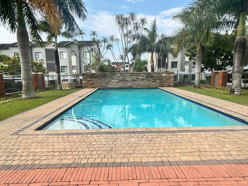 A well-maintained communal swimming pool area featuring a stone feature wall, brick paved surround, and established palm trees within an apartment complex.