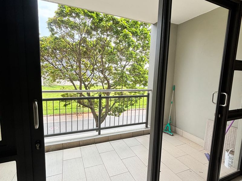 A clean, covered balcony featuring tiled flooring, a metal balustrade, and sliding glass door access, offering a pleasant view of the surrounding greenery.