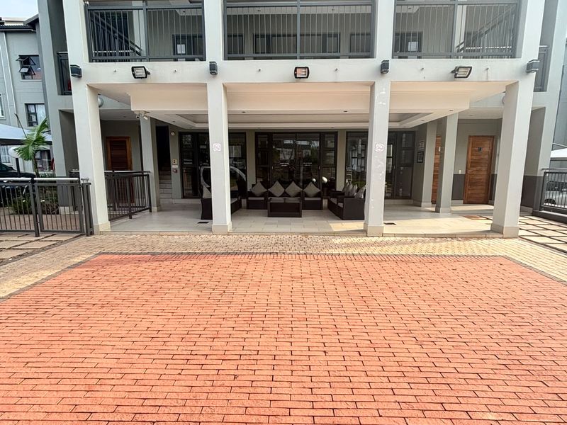 A well-maintained apartment building exterior featuring a paved driveway, covered ground-floor patio, and an upper-level balcony with metal railings.