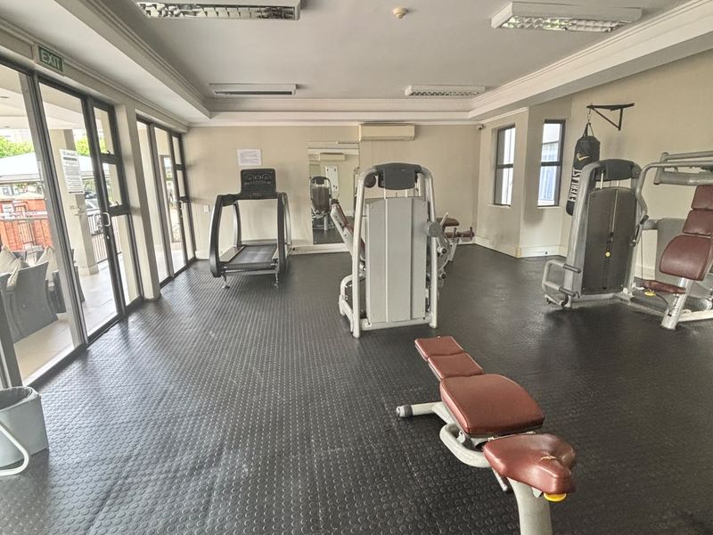 A functional apartment gym facility featuring rubber flooring, wall-mounted mirrors, air conditioning, and direct access to an outdoor patio area.