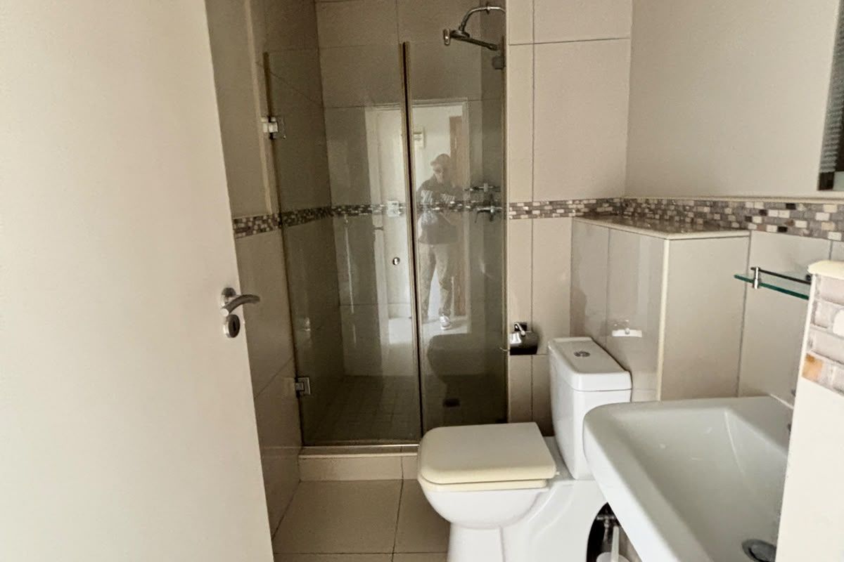A clean, functional bathroom featuring a glass-enclosed shower, pedestal basin, and neutral wall and floor tiling with a decorative mosaic accent.