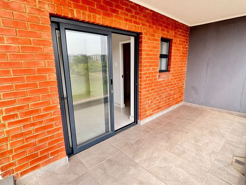 A clean, covered balcony featuring modern tiled flooring, an exposed brick feature wall, and a sliding glass door providing access to the interior.