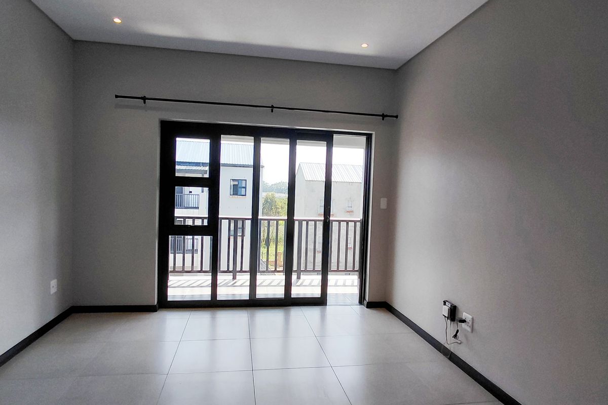 A bright, empty living room featuring modern tiled flooring, recessed lighting, and a large sliding glass door providing access to a balcony.