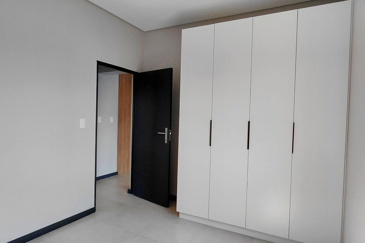 A clean, modern bedroom featuring neutral tiled flooring, recessed ceiling lighting, and a large white built-in wardrobe with dark handles.