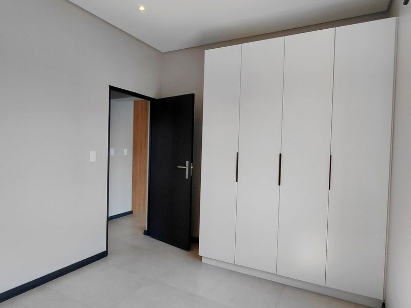 A clean, modern bedroom featuring neutral tiled flooring, recessed ceiling lighting, and a large white built-in wardrobe with dark handles.