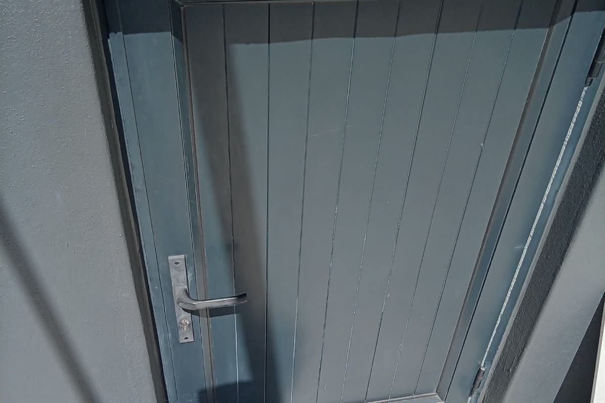 A close-up view of a grey exterior door with a metal handle, set against a matching grey wall and light-colored tiled flooring.