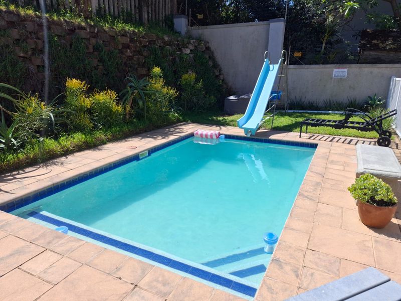 Outdoor swimming pool area featuring a built-in slide, paved surround, and a stone retaining wall in a landscaped garden setting.