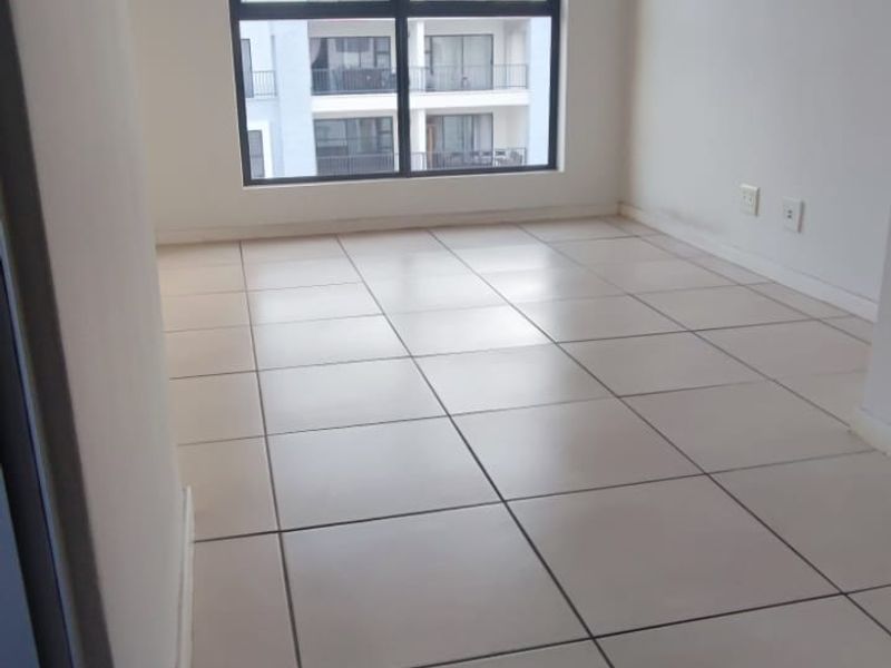 A bright, empty bedroom featuring neutral tiled flooring, a large window providing natural light, and a simple pendant light fixture.