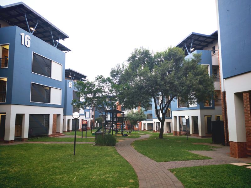 A well-maintained communal garden area between multi-story apartment blocks, featuring paved walkways, mature trees, and modern blue and brick exterior finishes.