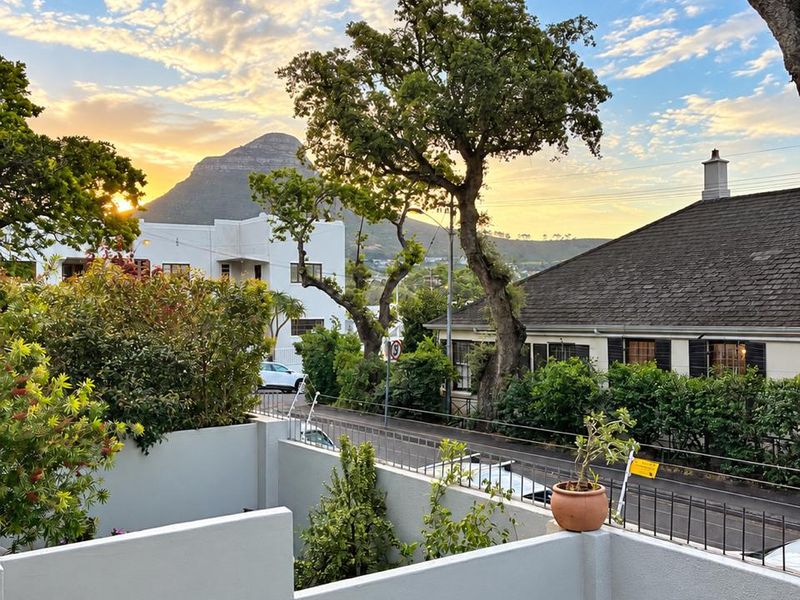 A scenic outdoor view from a balcony overlooking a quiet street, mature trees, and a prominent mountain peak at sunset.