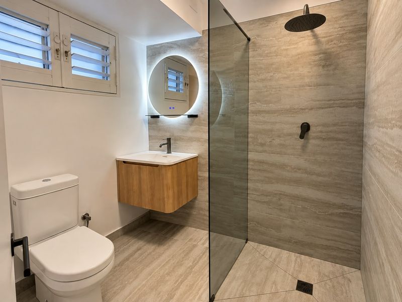Modern renovated bathroom featuring a walk-in shower with rain head, floating vanity, backlit mirror, and neutral stone-look tiling throughout.