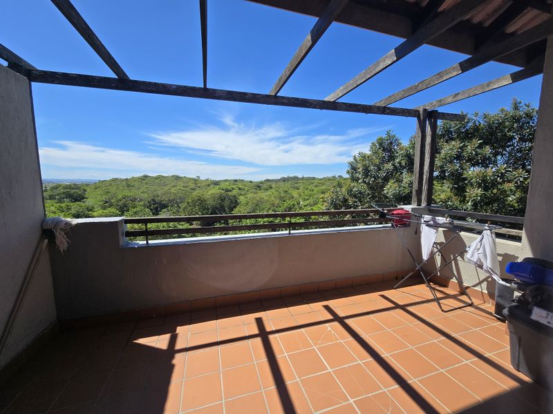A spacious balcony featuring terracotta floor tiles, a wooden pergola structure, and a metal railing, offering scenic views of the surrounding greenery.