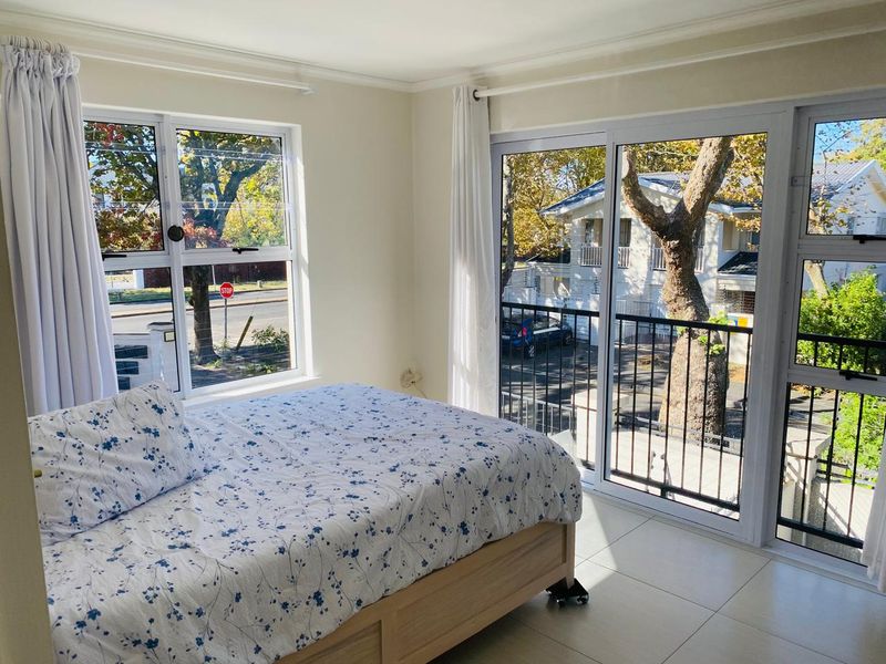 A bright bedroom featuring tiled flooring, large aluminium windows, and a sliding glass door leading to a private balcony.
