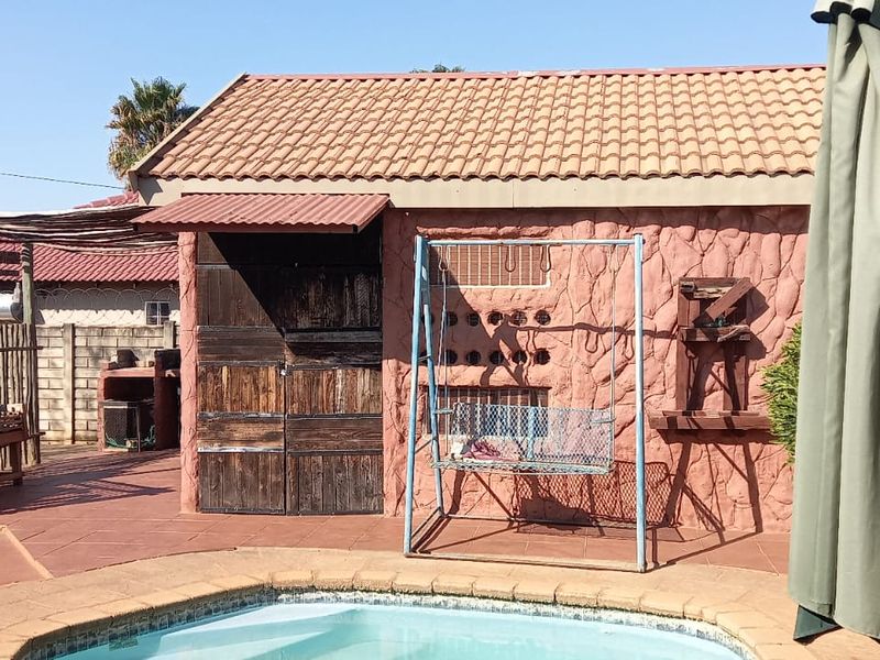 Outdoor pool area featuring a tiled patio, built-in braai, and a small outbuilding with a tiled roof, set within a fenced garden.