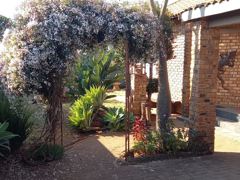 A covered patio area featuring exposed brickwork, sturdy brick pillars, and a paved floor, leading into a landscaped garden with a decorative metal archway.