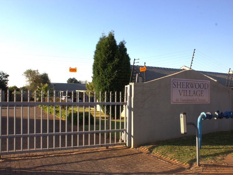 Front entrance view of Sherwood Village complex featuring a security gate, paved driveway, and perimeter boundary wall with electric fencing.