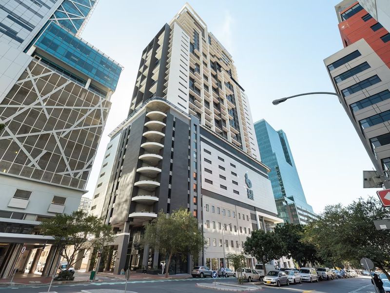 A modern high-rise apartment building featuring a prominent facade with multiple balconies, situated in a dense urban street environment.