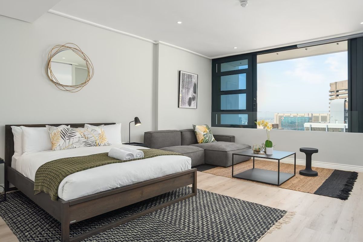 A modern, well-lit bedroom featuring light laminate flooring, recessed lighting, and a large sliding window offering expansive city views.