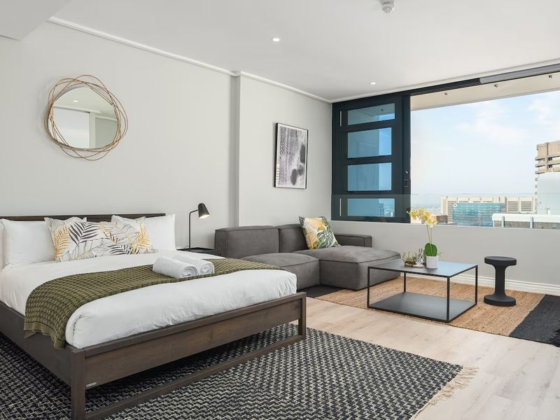 A modern, well-lit bedroom featuring light laminate flooring, recessed lighting, and a large sliding window offering expansive city views.
