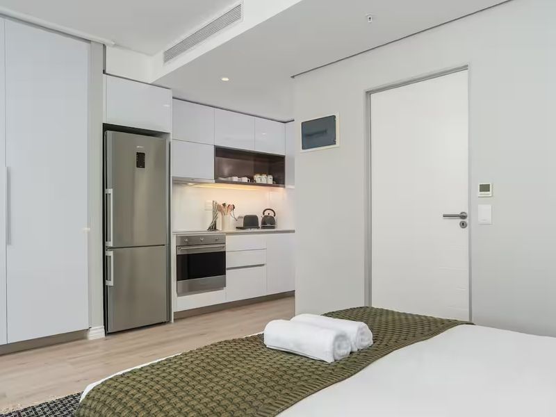 Modern open-plan studio apartment featuring a sleek kitchenette with integrated appliances, built-in cabinetry, and ducted air conditioning in a clean, contemporary finish.