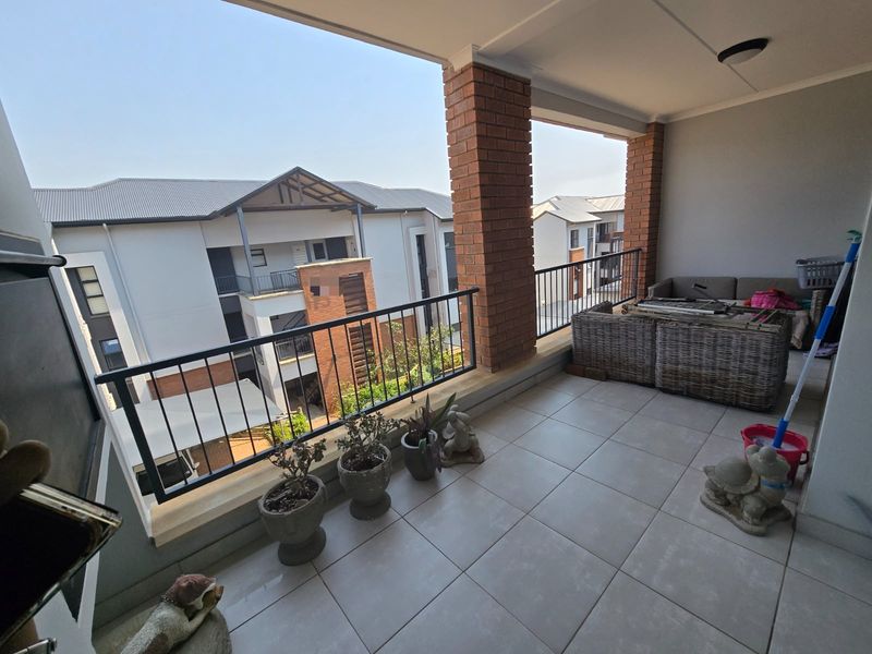 A spacious covered balcony featuring tiled flooring, brick support pillars, and a metal balustrade, offering an open view of the surrounding apartment complex.