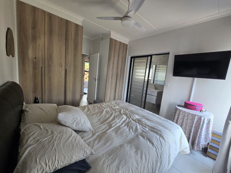 A well-maintained bedroom featuring extensive built-in wooden cupboards, a ceiling fan, and direct access to an en-suite bathroom with modern tiling.