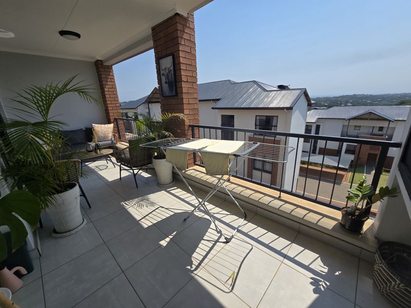 A spacious covered balcony featuring tiled flooring, a brick support pillar, and metal railings, offering an open view of the surrounding residential complex.