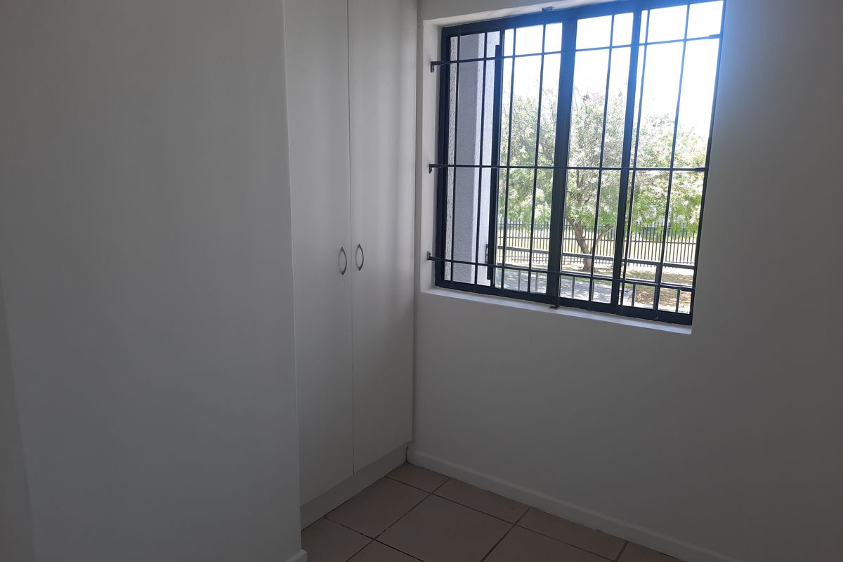 A clean, empty bedroom featuring neutral tiled flooring, built-in cupboards, and a window fitted with security bars.