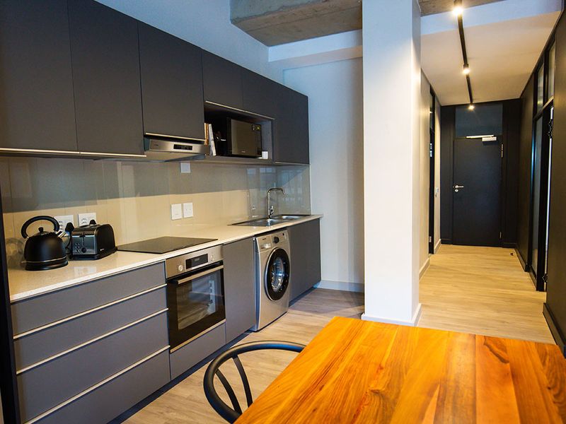 Modern kitchen featuring sleek dark cabinetry, stone countertops, integrated appliances, and an industrial-style exposed concrete ceiling, seamlessly flowing into the hallway.