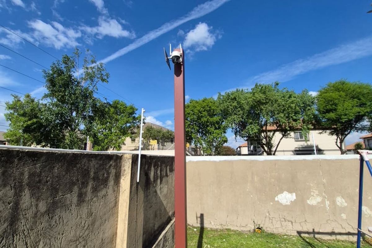 A rear outdoor area featuring a lawn, perimeter boundary wall with visible plaster damage, and a security camera mounted on a metal post.